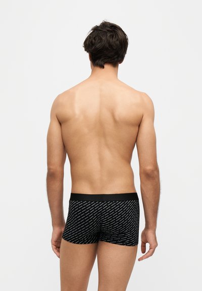 Homme debout dos à la caméra portant un boxer noir à motifs sur un fond clair uni.