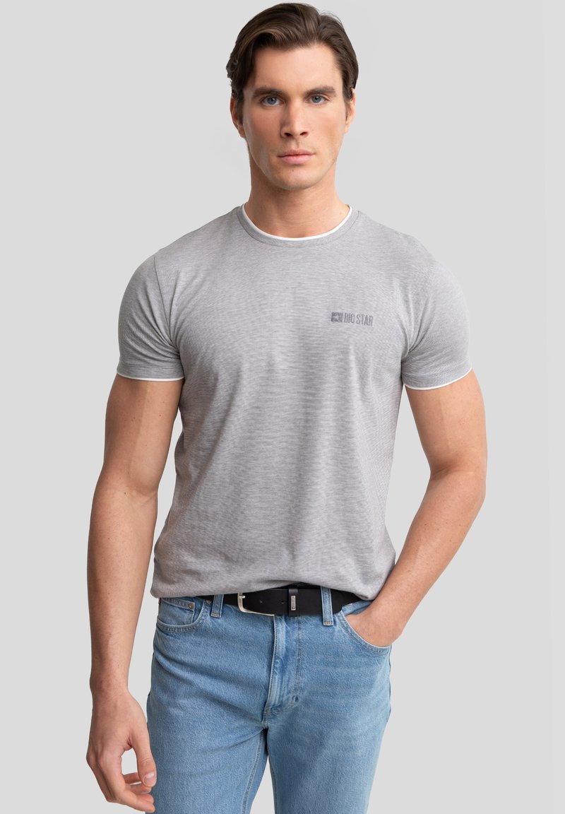 Camiseta gris de manga corta con un ribete blanco en el cuello, con un logo en el pecho, combinada con jeans de color azul claro y un cinturón negro.