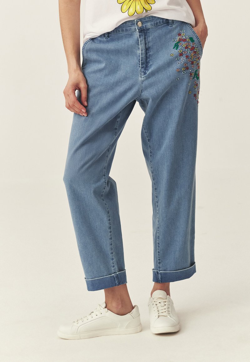 Frau trägt hellblaue Jeans mit Umschlag, verziert mit bunter Blumenstickerei auf der Tasche, und weiße Turnschuhe, steht vor einfachem Hintergrund.