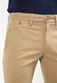 Béžové bavlněné chino kalhoty s hladkou texturou, jemným leskem, kontrastním prošíváním a malým modro-červeným vzorem na přední kapse.