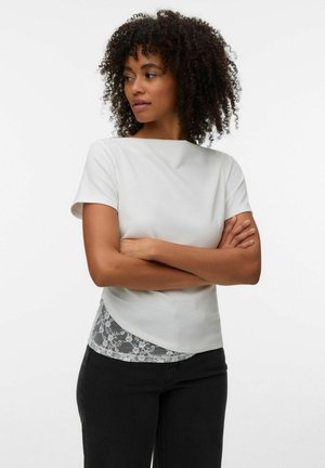 Femme aux cheveux bouclés portant un haut blanc à manches courtes par-dessus un caraco en dentelle et un pantalon noir, les bras croisés, regardant sur le côté.