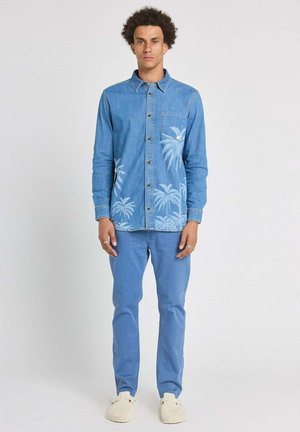Jeune homme debout, portant une chemise en denim bleu avec des imprimés de palmiers, un pantalon bleu et des chaussures blanches sans lacets, sur un fond blanc uni.