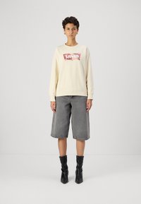 Krémová mikina s ružovým kvetinovým logom Levi's, v kombinácii so šedými džínsovými bermudami a čiernymi členkovými topánkami s riasenou textúrou.