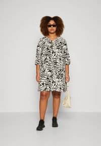 Black and white zebra-patterned dress with a V-neck, three-quarter sleeves, and relaxed fit. Accessories include a cream-colored bag and black shoes.