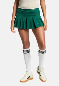 Zelená plisovaná sukňa s krátkym strihom a priliehavým pásom, s bielym logom a značkovým dizajnom "adidas". Nosená s bielymi ponožkami po kolená so čiernymi pruhmi.