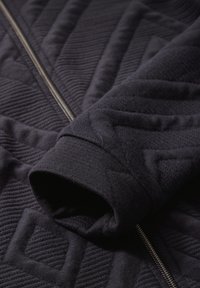 Veste matelassée noire avec un motif texturé, dotée d'une fermeture éclair argentée et de poignets côtelés, mettant en avant un tissu doux et chaud.