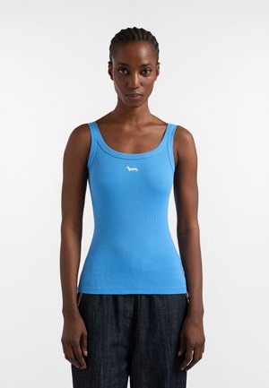 Donna con capelli intrecciati che indossa una canotta aderente blu con un piccolo logo bianco a forma di cane e pantaloni scuri, in piedi davanti a uno sfondo bianco uniforme.