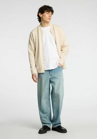 Beige bombázódzseki fehér póló fölött, világoskék, bő nadrággal és fekete, textúrázott mintájú cipőkkel párosítva.