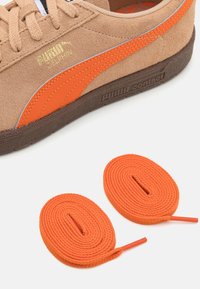 Baskets en daim beige avec une bande orange, associées à une semelle marron texturée et deux lacets plats orange placés sous la chaussure.