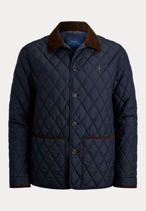 Homme portant des lunettes de soleil, une veste matelassée bleu marine, un pull en maille torsadée olive, un jean bleu clair, des bottes en daim marron, debout avec une main dans la poche.