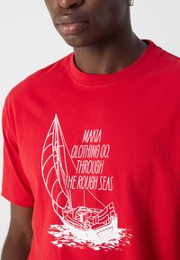 Red cotton t-shirt featuring a white graphic of a sailboat and text "MAKIA CLOTHING CO. THROUGH THE ROUGH SEAS" on the front.