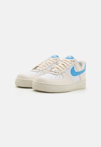 Krémové tenisky s koženým svrškem, modrým logem Nike, perforovaným špičkovým dílem, texturovanou mezipodešví a béžovou gumovou podrážkou.
