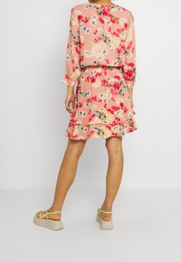 Květinové šaty v jemně růžové barvě s dlouhými rukávy, nařaseným pasem a volánkovým lemem, zdobené vzory červených a žlutých květin. Doplněné o béžové sandály na platformě.