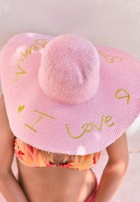 Širokorozsáklý růžový slaměný klobouk se žlutým vyšitým nápisem "I love" a "beach." Má texturované pletení a volný tvar.