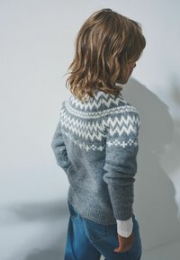 Maglione grigio a maglia con un motivo geometrico bianco sul giromanica, dotato di maniche lunghe e orlo a coste, indossato sopra una camicia bianca.