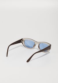 Óculos de sol transparentes com detalhes em castanho e lentes azuis, apresentando uma forma distinta de olho de gato e um detalhe de logotipo na haste.