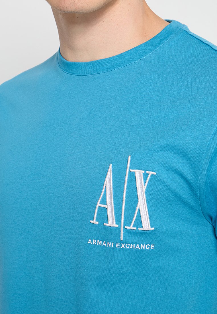 Mężczyzna nosi jasnoniebieską koszulkę z krótkim rękawem, z białym haftowanym logo "A|X Armani Exchange" na piersi, przyciętą poniżej nosa.