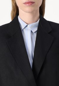 Blazer noir avec une finition texturée, superposé à une chemise boutonnée à rayures bleu clair. Le col de la chemise est visible sous le blazer.