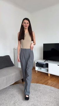 Γυναίκα με μακριά καστανά μαλλιά που φοράει μπεζ αμάνικη μπλούζα και γκρι παντελόνι περπατάει σε έναν στρογγυλό μπεζ χαλί σε ένα σαλόνι.