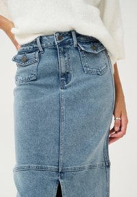 Jeansrock in Hellblau mit einem Schlitz vorne, großen aufgesetzten Taschen, sichtbaren Nähten und Metallknopf-Details. Getragen mit einem weißen, strukturierten Pullover.