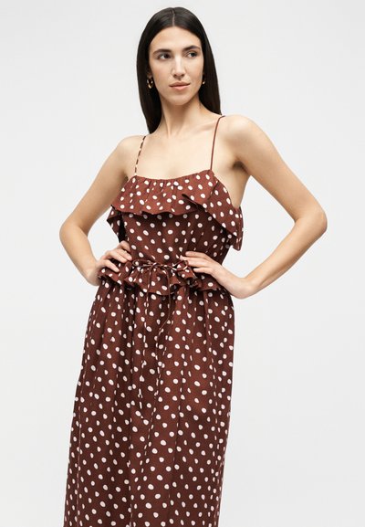 Γυναίκα με καφέ αμάνικο φόρεμα με λευκές πουά και λεπτομέρειες με βολάν, στέκεται με χέρια στη μέση πάνω σε απλό φόντο.