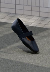 Črne usnjene ravne čevlje z zaobljenim nosom, ki imajo mehko črno elastično naramnico čez vamp. Gladka tekstura in minimalističen dizajn.