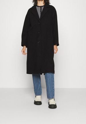 Persona che indossa un lungo cappotto nero sopra una maglia a righe, jeans blu e stivaletti bianchi con suola nera, in piedi davanti a uno sfondo bianco semplice.