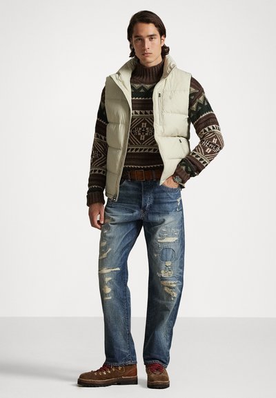 Colete acolchoado em creme claro com um bolso zippado, suéter de malha castanho estampado, jeans azul desgastados e botas castanhas com atacadores.