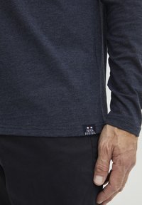 Temno moderna majica z dolgimi rokavi iz teksturirane mešanice bombaža, s majhno oznako blagovne znamke blizu roba in fitted manšeti. Gladek zaključek tkanine.