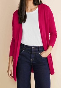 Fuchsieový kardigan s dlouhými rukávy, otevřený design, lehká pletenina, kombinovaný s bílým tričkem a tmavě modrými džíny.