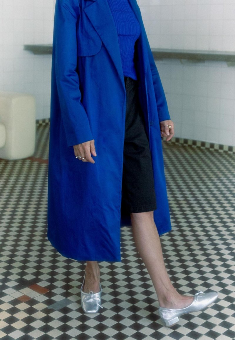 Cappotto blu, top blu a coste, pantaloni corti neri e scarpe argento con fiocco. Il pavimento presenta un motivo a piastrelle in bianco e nero.
