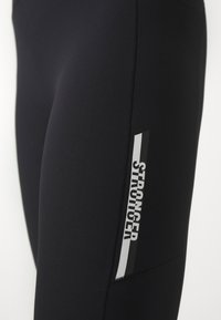 Fekete kompressziós leggings sima textúrával, az oldalon függőleges "STRONGER" logóval, fehér és szürke részletekkel.