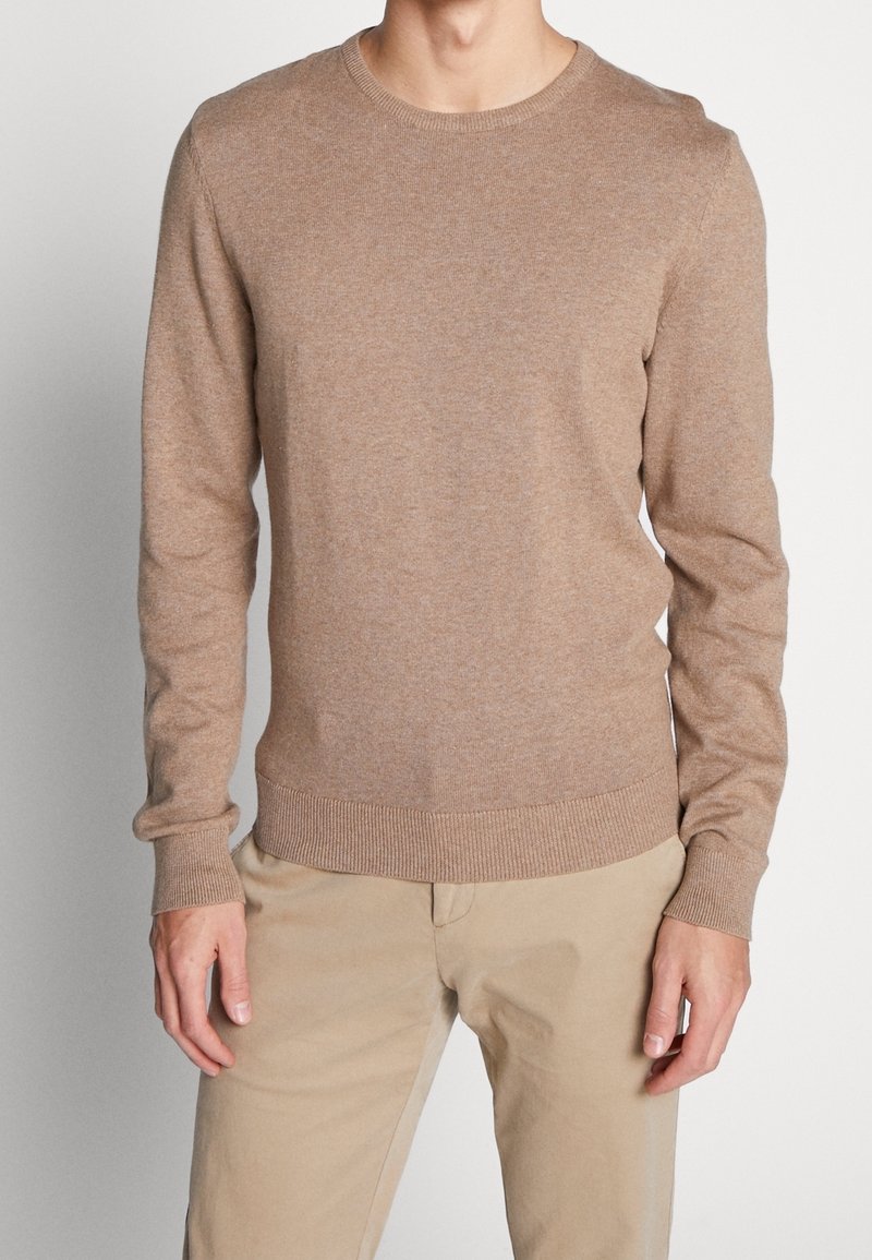 Maglione in maglia marrone con collo rotondo, maniche lunghe e polsini e orlo a costine. Tessuto morbido, vestibilità casual, indossato con pantaloni beige chiaro.