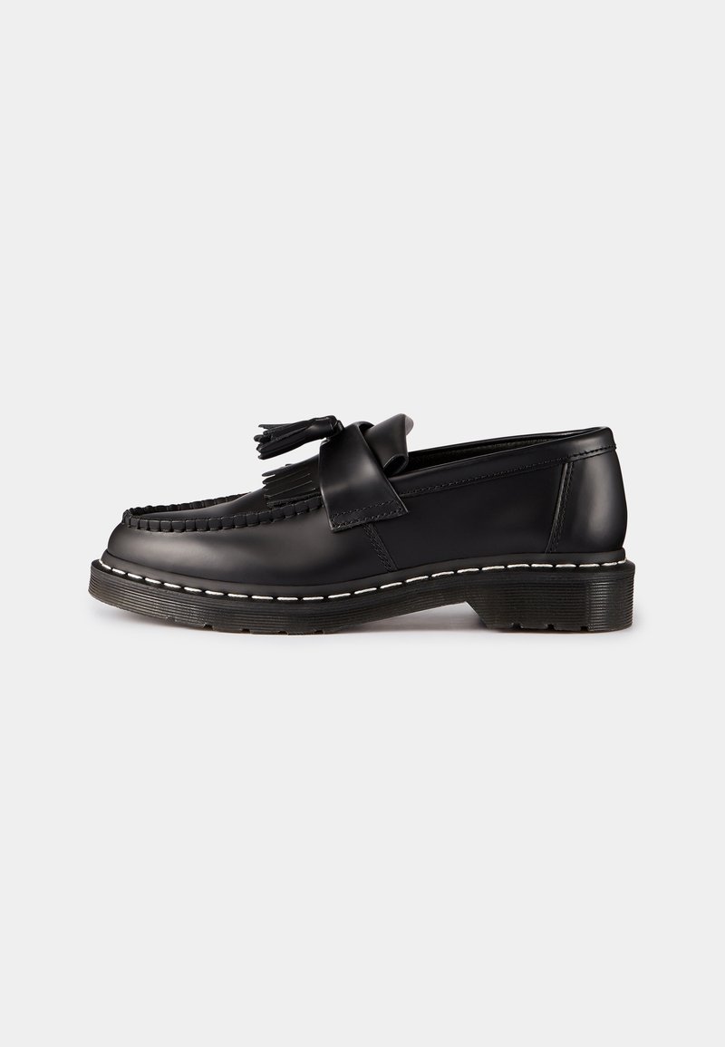 Mocassin en cuir noir avec glands, détail à franges, couture blanche sur semelle épaisse, montré de côté sur un fond clair uni.