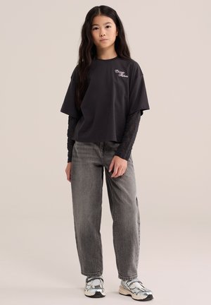Une jeune fille pose avec un T-shirt noir superposé, un jean gris et des baskets blanches sur un fond de studio beige uni.