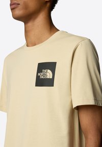 Beige tričko s krátkým rukávem vyrobené z měkké látky, s černým čtvercovým logem a nápisem "THE NORTH FACE" v bílém na levé straně hrudi.