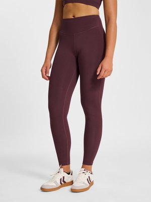 Vrouw die donkerpaarse legging met hoge taille en witte sneakers met bordeaux strepen draagt, staand tegen een effen achtergrond.