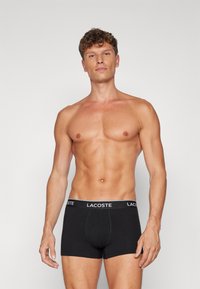 Crne pamučne bokserice s bijelim Lacoste logom na pojasu. Glatka tekstura i uski dizajn. Model ima definiranu tjelesnu građu.