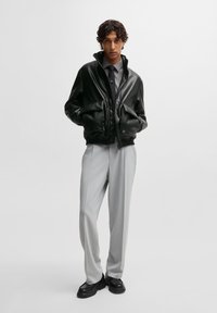 Svart lær bomberjakke med høy krage, frontglidelås og to store lommer, kombinert med lysegrå skreddersydde bukser og svarte sko.