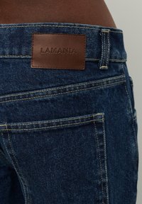 Tumedat sinised denim teksad, millel on taga vöökoha juures pruun nahkpook "LAMANIA", ja traditsioonilised tasku õmblusdetailid.