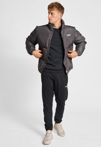 Giubbotto piumino grigio con colletto alto, chiusura a zip e polsini a coste, indossato sopra una maglietta nera a girocollo e pantaloni della tuta neri, con sneakers beige.