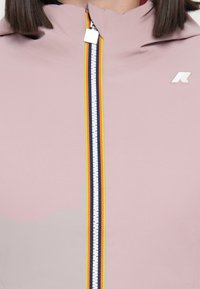 Light pink waterproof jacket with a high collar, featuring a contrasting zipper in white, navy, and yellow colors. Includes logo detail.