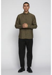 Olivno zelena košulja s dugim rukavima, s teksturiranom tkaninom, crnim gumbima i zakrivljenim rubom, u kombinaciji s crnim hlačama i cipelama.
