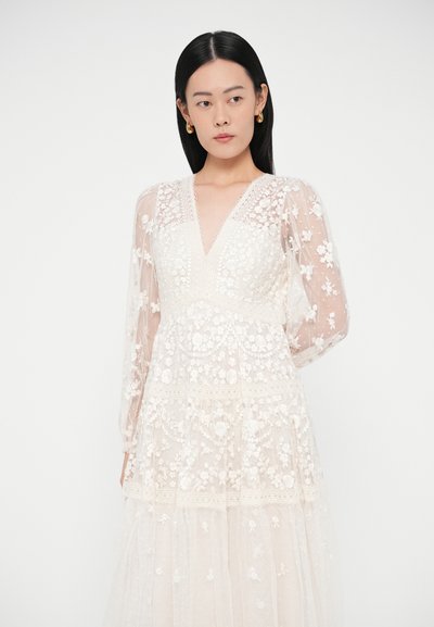 Femeie cu păr negru lung, purtând o rochie albă din dantelă cu flori, cu mâneci lungi transparente și cercei aurii în formă de cerc, stând în fața unui fundal alb.