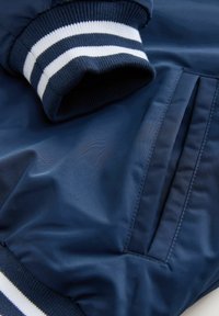 Marineblaues Bomberjackett mit gerippten Bündchen mit weißen Streifen, glatter Textur und seitlichem Taschen-Detail.