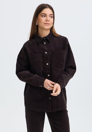 Femme aux longs cheveux bruns porte une chemise sombre ample à boutons et un pantalon assorti, regarde sur le côté, mains jointes.