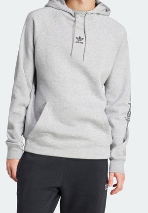 Osoba nosząca jasnoszarą bluzę z kapturem marki Adidas z przednią kieszenią i logo na piersi, w zestawieniu z czarnymi spodniami Adidas z logo na udzie.