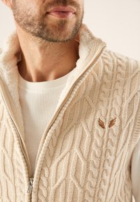 Gilet en tricot beige avec un col haut, présentant un motif en côtes et une doublure en polaire douce. Comprend un logo décoratif en marron.