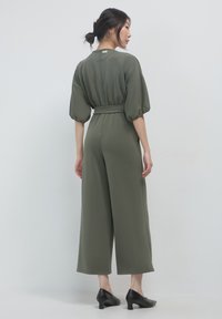 Combinaison verde oliva con maniche corte e puff; vita cinchata con un nastro; design con gambe larghe; texture liscia; indossata con scarpe nere con tacco.