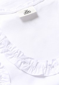 Top de algodón blanco con textura acanalada y un cuello con volantes. La etiqueta con el logo aparece en la parte posterior del cuello.